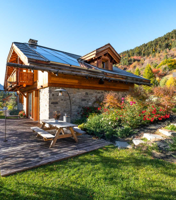 Un Chalet de montagne avec sa terrasse et une table de pique-nique en bois en bois avec une pelouse verte au premier plan